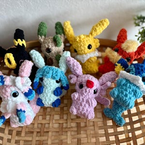 May include: A selection of handmade crochet plush toys in various colours, including yellow, red, blue, and pink. The toys are arranged in a woven basket and feature large eyes and animal-like features.