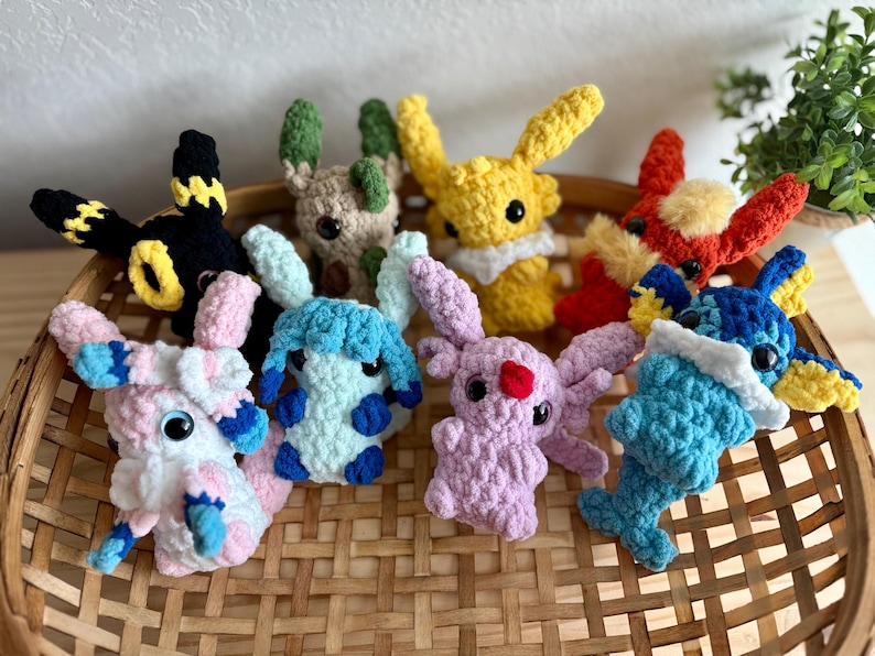 May include: A selection of handmade crochet plush toys in various colours, including yellow, red, blue, and pink. The toys are arranged in a woven basket and feature large eyes and animal-like features.