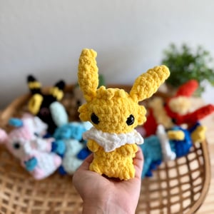 May include: A yellow crochet plush toy with large ears and a white collar is held in a hand. Other colourful crochet toys are visible in a woven basket in the background. The plush toy has black eyes.