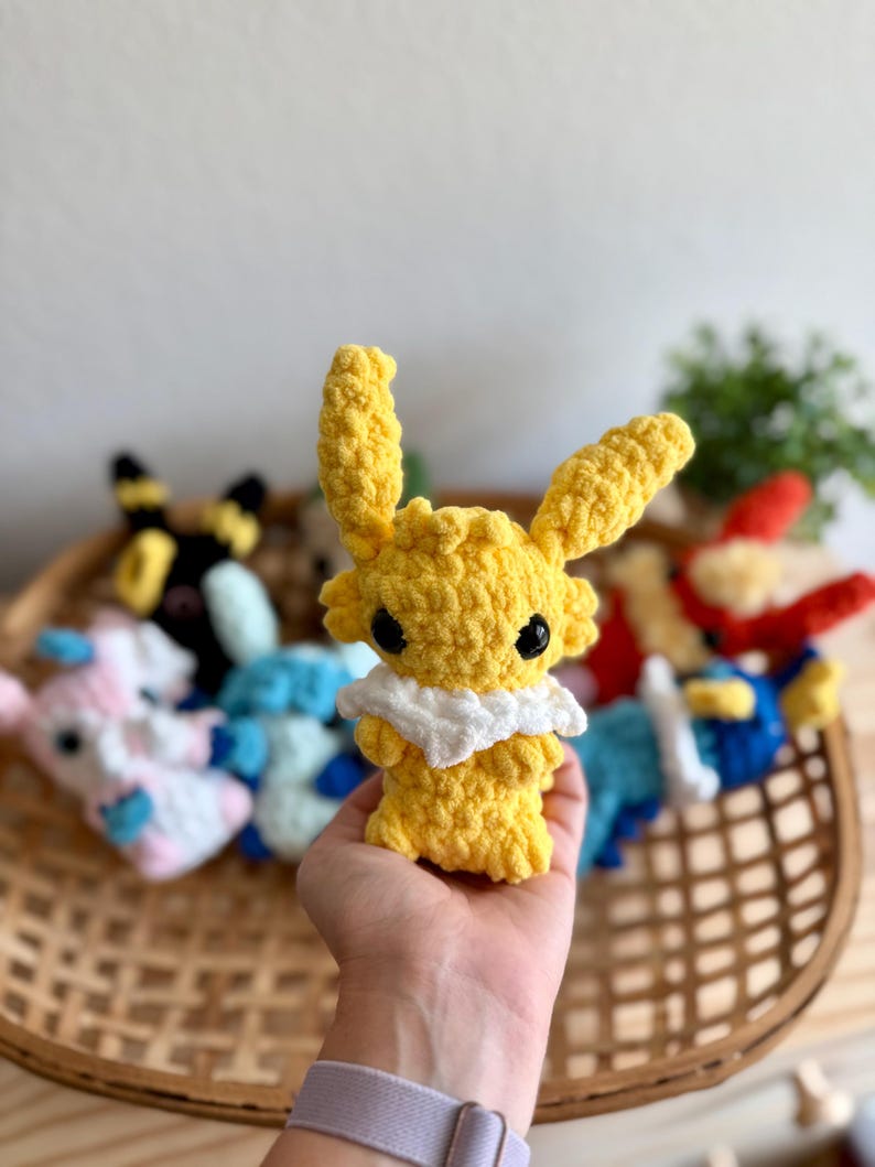 May include: A yellow crochet plush toy with large ears and a white collar is held in a hand. Other colourful crochet toys are visible in a woven basket in the background. The plush toy has black eyes.