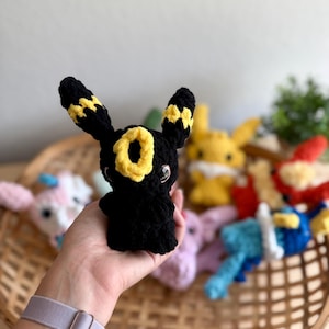 May include: A black crochet Umbreon plushie with yellow detailing, held in a hand. The plushie features large ears and a yellow ring on its forehead. Other colourful crochet figures are in the background, including Pikachu and other Pok&eacute;mon.