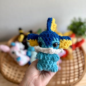 May include: A handmade crochet plush toy resembling a cartoon character. The toy is mainly blue with yellow detailing on the ears and head. It features black eyes and a white collar. Other similar toys are visible in a wicker basket in the background.