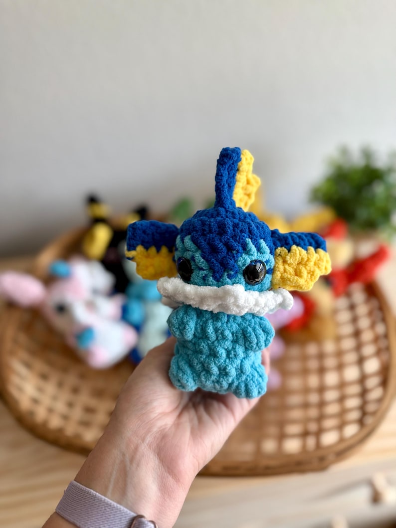 May include: A handmade crochet plush toy resembling a cartoon character. The toy is mainly blue with yellow detailing on the ears and head. It features black eyes and a white collar. Other similar toys are visible in a wicker basket in the background.