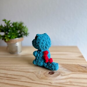 K&ouml;nnte beinhalten: Ein tealfarbenes geh&auml;keltes Dinosaurierspielzeug mit schwarzen Augen und roten Akzenten auf R&uuml;cken und Schwanz. Das handgefertigte Kuscheltier sitzt auf einer Holzoberfl&auml;che. Eine kleine Topfpflanze befindet sich im Hintergrund.