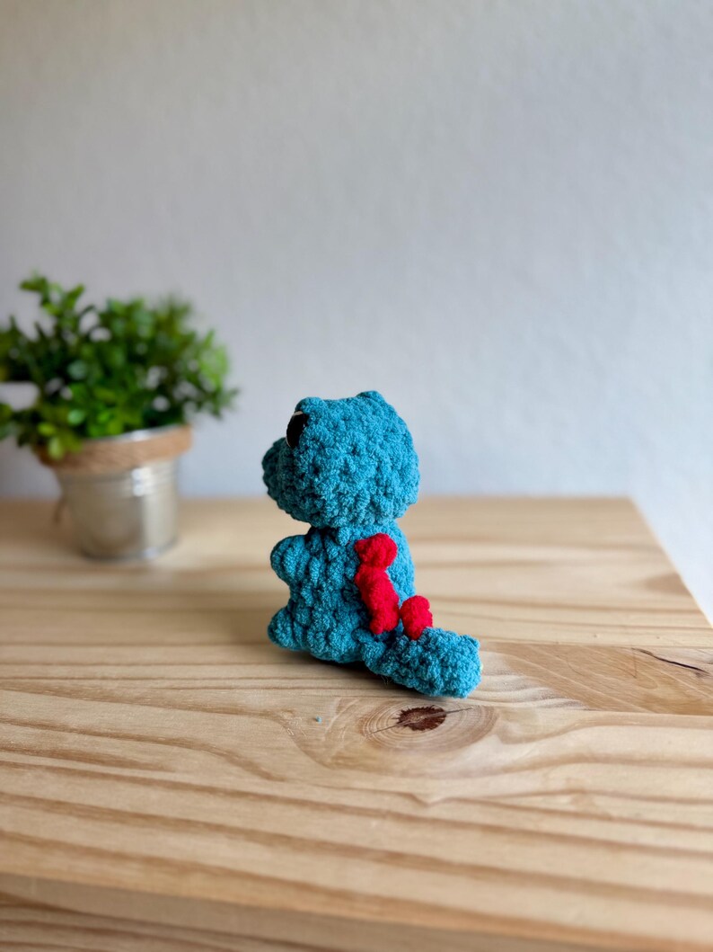 K&ouml;nnte beinhalten: Ein tealfarbenes geh&auml;keltes Dinosaurierspielzeug mit schwarzen Augen und roten Akzenten auf R&uuml;cken und Schwanz. Das handgefertigte Kuscheltier sitzt auf einer Holzoberfl&auml;che. Eine kleine Topfpflanze befindet sich im Hintergrund.
