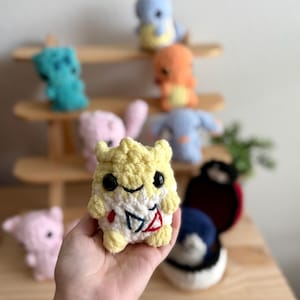 May include: A yellow and white crocheted plush toy with a red heart and a smiling face. The toy is being held in a person's hand. There are other crocheted plush toys in the background.