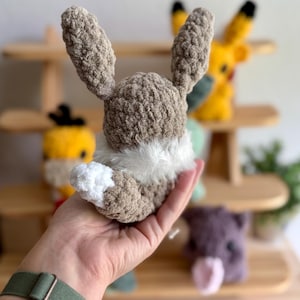 Puede incluir: Un peluche de conejo hecho a mano con ganchillo en tonos marr&oacute;n, blanco y beige. El conejo tiene orejas largas y un cuello blanco y esponjoso. Otros juguetes de ganchillo son visibles en el fondo, incluido un Pikachu amarillo.