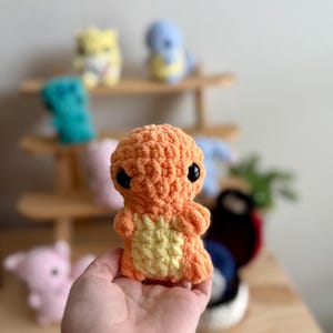 Könnte beinhalten: Ein gehäkeltes Plüschtier in Orange und Gelb, das einem Pokemon-Charakter ähnelt. Das Spielzeug hat schwarze Augen und einen kleinen Mund. Das Spielzeug wird in der Hand einer Person gehalten.