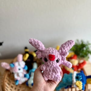 May include: A lavender-coloured crochet creature with large ears and a red heart-shaped nose. The plush toy is held in a hand, with other colourful crochet toys visible in a woven basket in the background. The toys are a variety of colours.