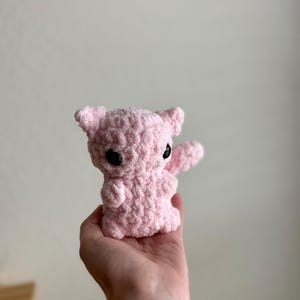 May include: A pink crocheted stuffed animal with black eyes. The animal is being held in a person's hand.