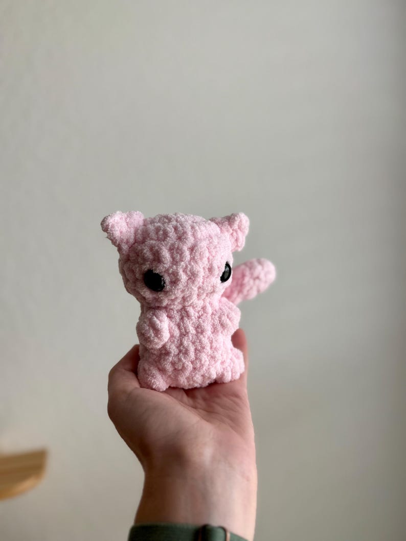May include: A pink crocheted stuffed animal with black eyes. The animal is being held in a person's hand.