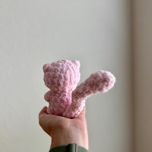 May include: A pink crocheted stuffed animal in the shape of a cat with a long tail. The cat is being held by a hand.