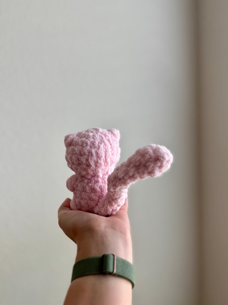 May include: A pink crocheted stuffed animal in the shape of a cat with a long tail. The cat is being held by a hand.