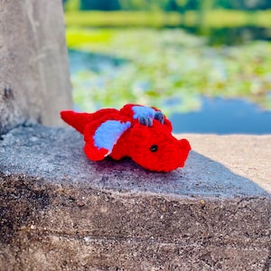 Pu&ograve; includere: Un piccolo drago rosso all'uncinetto con ali blu. Il drago &egrave; sdraiato su una superficie di cemento grigia.