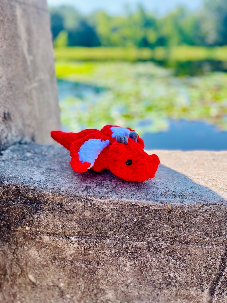 Pu&ograve; includere: Un piccolo drago rosso all'uncinetto con ali blu. Il drago &egrave; sdraiato su una superficie di cemento grigia.
