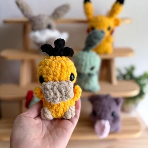 May include: A crocheted yellow Psyduck plush toy with a black headpiece and a tan beak and feet. Other crocheted plush toys are visible in the background, including a Pikachu, a bunny, a green creature, and a purple pig.