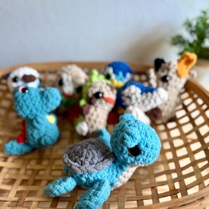 Puede incluir: Una colección de animales de ganchillo hechos a mano, incluyendo una tortuga azul con caparazón gris, un dinosaurio azul y varias otras criaturas. Los juguetes se exhiben en una cesta tejida, mostrando sus texturas suaves y colores vibrantes.