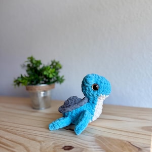 Puede incluir: Una tortuga de ganchillo hecha a mano en tonos de azul, gris y blanco. La tortuga tiene un aspecto texturizado y afelpado con una cabeza redondeada, ojos pequeños y un caparazón gris. Está colocada sobre una superficie de madera.