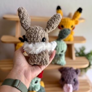 May include: A crocheted brown bunny with white fur around its neck is held in a hand. Other crocheted animals, including a yellow creature with black-tipped ears, are visible on a wooden shelf. The bunny has black eyes.