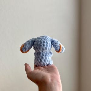 May include: A small, light blue crocheted bunny with orange detailing on the ears. The handmade plush toy is held in a hand, highlighting its soft texture and craftsmanship. The bunny is a cute, handmade item, perfect for a child.