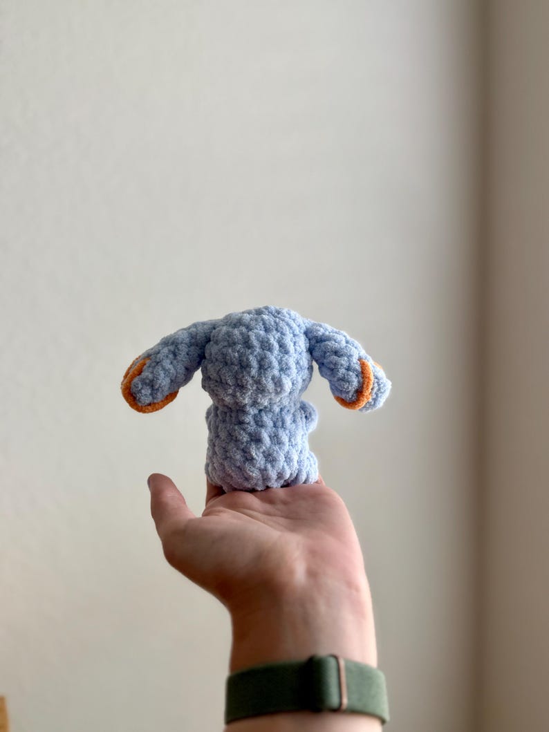 May include: A small, light blue crocheted bunny with orange detailing on the ears. The handmade plush toy is held in a hand, highlighting its soft texture and craftsmanship. The bunny is a cute, handmade item, perfect for a child.