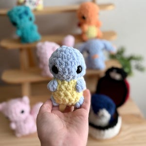 Puede incluir: Un personaje de Pokemon de crochet azul y amarillo con ojos negros. El personaje está siendo sostenido en la mano de una persona.
