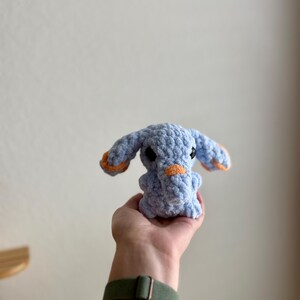 May include: A handmade, light blue crochet elephant with orange detailing on the ears and trunk. The soft toy has small black eyes. The elephant is held in a hand. A cuddly toy.