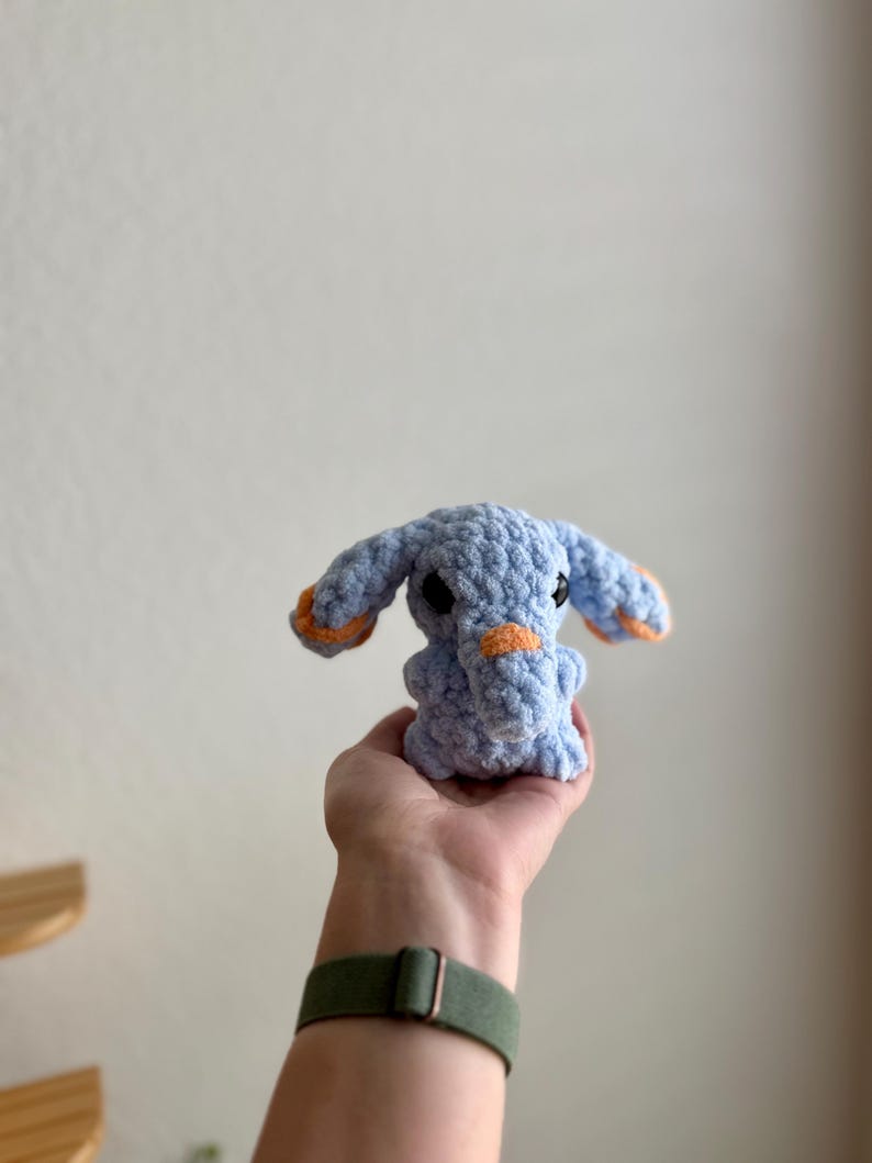May include: A handmade, light blue crochet elephant with orange detailing on the ears and trunk. The soft toy has small black eyes. The elephant is held in a hand. A cuddly toy.