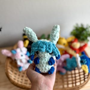 May include: A handmade crochet plush toy in the shape of a creature with light blue and white yarn. The toy has large ears, blue accents, and is held in a hand. Other similar toys are in a basket in the background.
