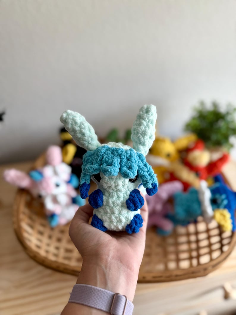 May include: A handmade crochet plush toy in the shape of a creature with light blue and white yarn. The toy has large ears, blue accents, and is held in a hand. Other similar toys are in a basket in the background.