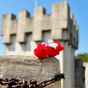 Pu&ograve; includere: Un drago rosso all'uncinetto con ali bianche &egrave; seduto su un muro di pietra con una catena arrugginita. Il drago &egrave; rivolto verso la fotocamera e ha un piccolo naso bianco. Il muro &egrave; fatto di pietra grezza e ha una consistenza ruvida. La catena &egrave; arrugginita e mancano alcuni anelli.