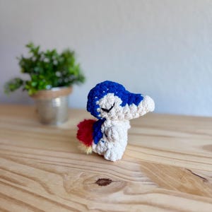 Op de afbeelding: Een handgemaakte gehaakte figuur van een klein, gestileerd wezen. Het heeft een blauwe kop met witte accenten, een crèmekleurig lichaam en een rood-gele staart. De figuur staat op een houten oppervlak.