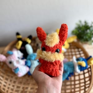 May include: A handmade, red and yellow crocheted plush toy with large black eyes and upright ears. The plush is held in a hand, with other colourful plush toys in the background, all inside a woven basket.