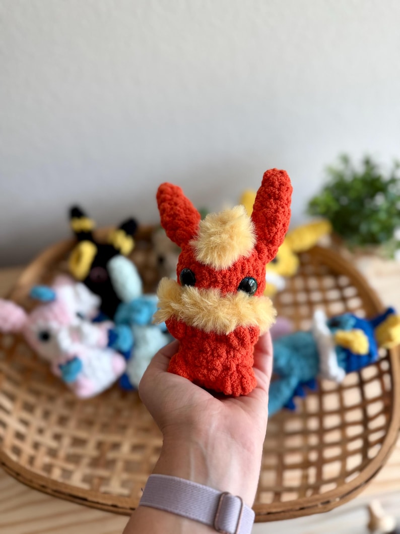 May include: A handmade, red and yellow crocheted plush toy with large black eyes and upright ears. The plush is held in a hand, with other colourful plush toys in the background, all inside a woven basket.
