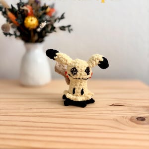 May include: A cream-colored crochet plush toy with black ear tips, black feet, and a black stitched face featuring a star-shaped eye and a wide mouth. The figure has red accents on the sides of its head and the text Tester call open in yellow.