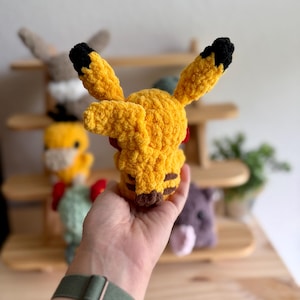 May include: A handmade yellow Pikachu plush toy with black-tipped ears and brown stripes on the cheeks and back. The plush is made of a soft, textured yarn. Other plush toys are visible in the background on a wooden shelf.