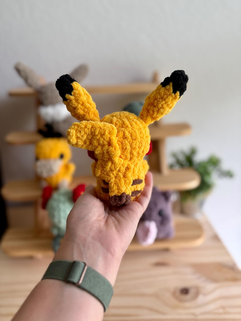May include: A handmade yellow Pikachu plush toy with black-tipped ears and brown stripes on the cheeks and back. The plush is made of a soft, textured yarn. Other plush toys are visible in the background on a wooden shelf.