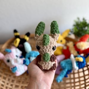 May include: A crocheted plush toy shaped like a creature with large eyes, green ears, and brown details. The toy is held in a hand, with other colourful crocheted toys in a basket in the background.
