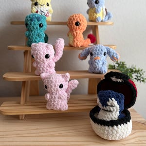 May include: A collection of crocheted Pokemon characters, including Pikachu, Squirtle, Charmander, Bulbasaur, and more. The characters are displayed on a wooden shelf.