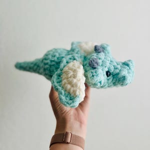 Pu&ograve; includere: Un giocattolo di dinosauro all'uncinetto color turchese con dettagli bianchi e grigi. Il dinosauro ha una coda lunga e ali piccole e arrotondate.