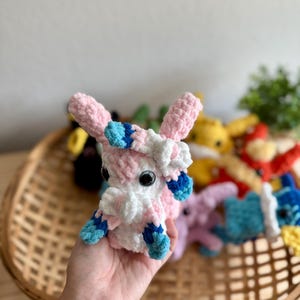May include: A handmade crochet plush toy in pink, white, and blue. The plush has large black eyes and is held in a wicker basket with other colourful crochet toys. The toy is approximately 10 cm tall.