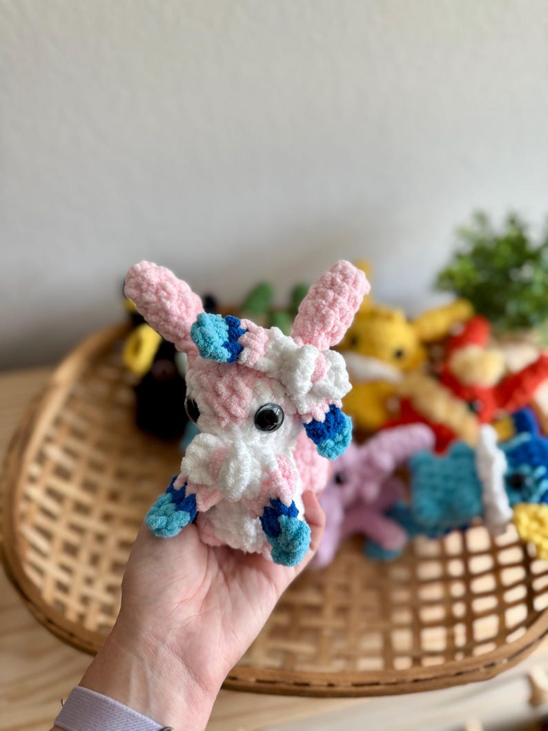 May include: A handmade crochet plush toy in pink, white, and blue. The plush has large black eyes and is held in a wicker basket with other colourful crochet toys. The toy is approximately 10 cm tall.