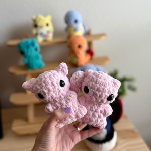 May include: Two pink crocheted plushie toys with black eyes. The plushies are being held in a person's hand.