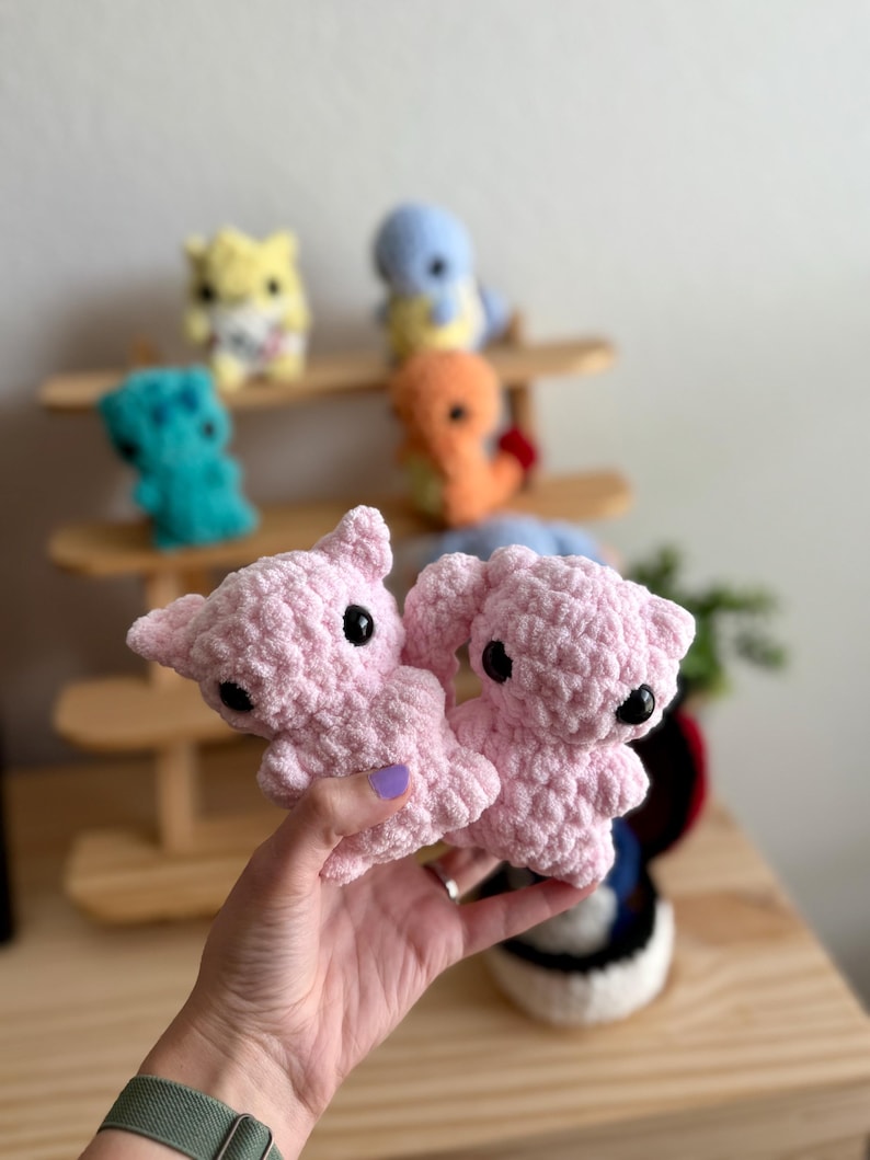 May include: Two pink crocheted plushie toys with black eyes. The plushies are being held in a person's hand.