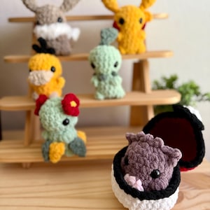 May include: A collection of handmade crochet Pokémon figures in various colors. The figures include Pikachu, a brown rabbit-like creature, a green creature, a yellow duck-like figure, a green and yellow creature with a red flower, and a purple creature in a Poké Ball.