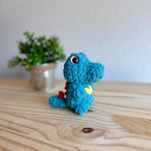 K&ouml;nnte beinhalten: Ein t&uuml;rkisfarbenes geh&auml;keltes Dinosaurierspielzeug mit rosa Augen, einem gelben Herz und roten Akzenten. Das handgefertigte Pl&uuml;schtier sitzt auf einer Holzoberfl&auml;che. Eine kleine Topfpflanze befindet sich im Hintergrund.
