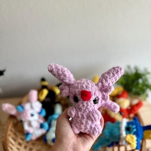 May include: A lavender-coloured crochet creature with large ears and a red heart-shaped nose. The plush toy is held in a hand, with other colourful crochet toys visible in a woven basket in the background. The toys are a variety of colours.