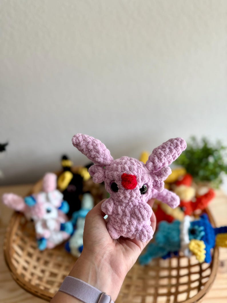 May include: A lavender-coloured crochet creature with large ears and a red heart-shaped nose. The plush toy is held in a hand, with other colourful crochet toys visible in a woven basket in the background. The toys are a variety of colours.