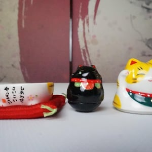 以下が含まれることがあります： 赤いクッションの上に、3つの日本の招き猫の置物と小さなボウルがあります。置物は黒、黄色、白で、様々なデザインが施されています。ボウルには日本語のテキストと花の装飾が施されています。
