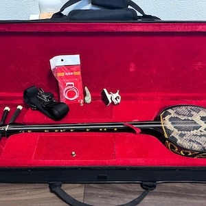 Könnte beinhalten: Ein Sanshin, ein dreisaitiges japanisches Musikinstrument mit einem mit Schlangenhaut bespannten Korpus, in einem schwarzen Hartschalenkoffer mit leuchtend rotem Samtinnenfutter. Enthalten sind ein schwarzer Gurt, ein weißes Stimmgerät und Zubehör.